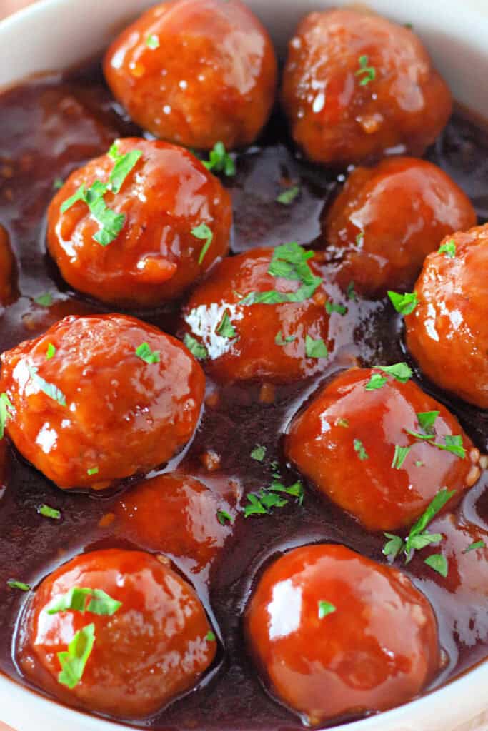A bowl of meatballs covered in a glossy brown sauce, garnished with chopped fresh herbs.