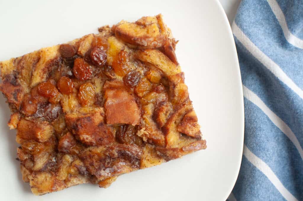 Overhead shot of a slice of cinnamon roll casserole with visible cinnamon swirls and raisins on a white plate.