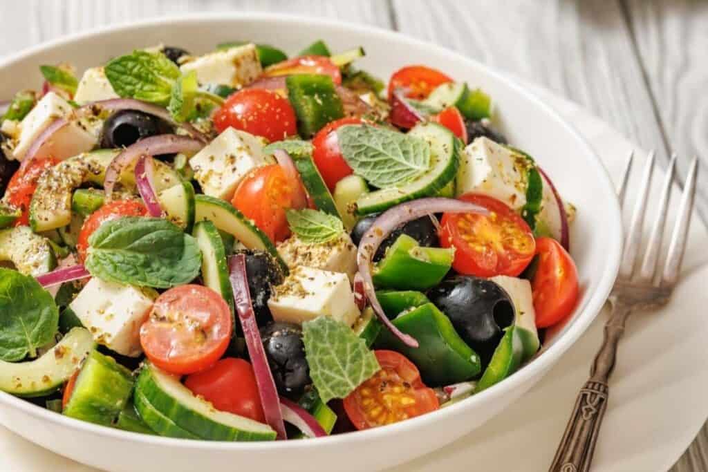 A bowl of Greek salad with cherry tomatoes, feta cheese, black olives, cucumber, red onion, green bell pepper, and fresh mint leaves. Forks are placed beside the bowl.