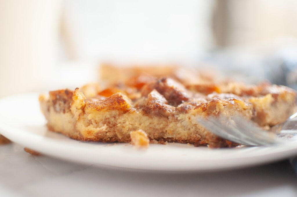 A close-up of a slice of golden-brown cinnamon roll casserole on a white plate with a fork.