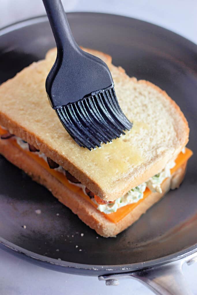 A pastry brush applies melted butter to the top slice of a stacked sandwich with cheese and other fillings in a skillet.