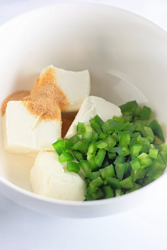 A white bowl containing three blocks of cream cheese, chopped green bell pepper, and a sprinkle of powdered seasoning.