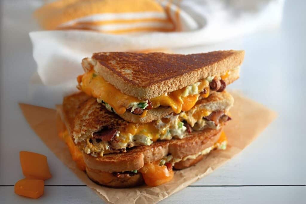 Two grilled cheese sandwiches with melted cheddar and fillings are stacked on parchment paper, with cheese cubes nearby and a folded cloth in the background.