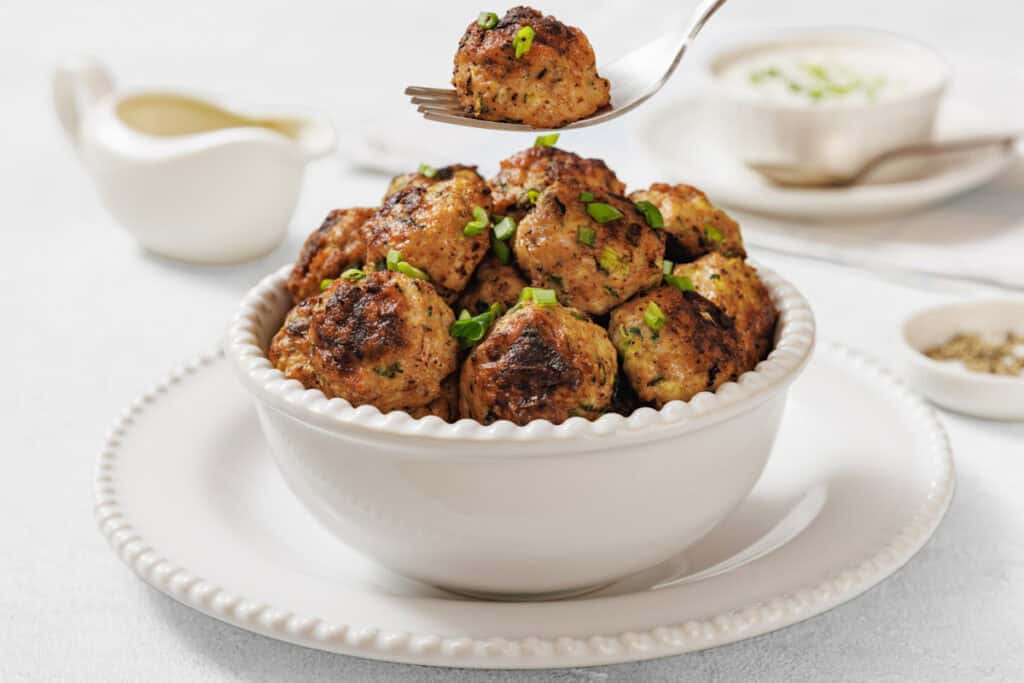 A bowl of cooked meatballs garnished with chopped herbs sits on a white plate, with a fork lifting one meatball above the bowl.