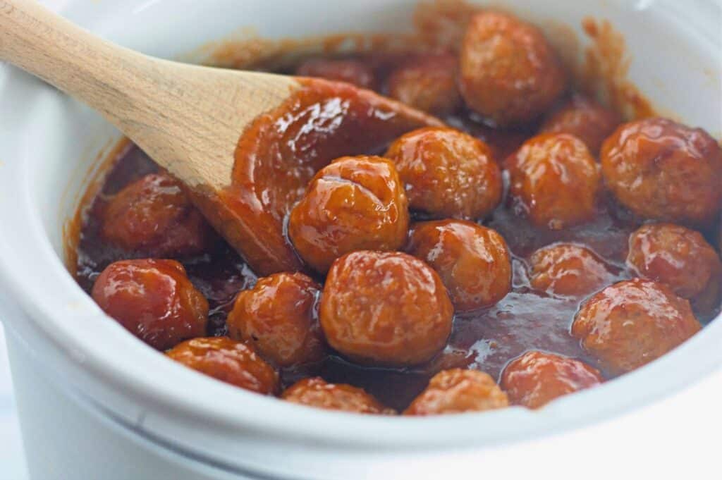 Meatballs in a thick red sauce being stirred with a wooden spoon in a white ceramic pot.