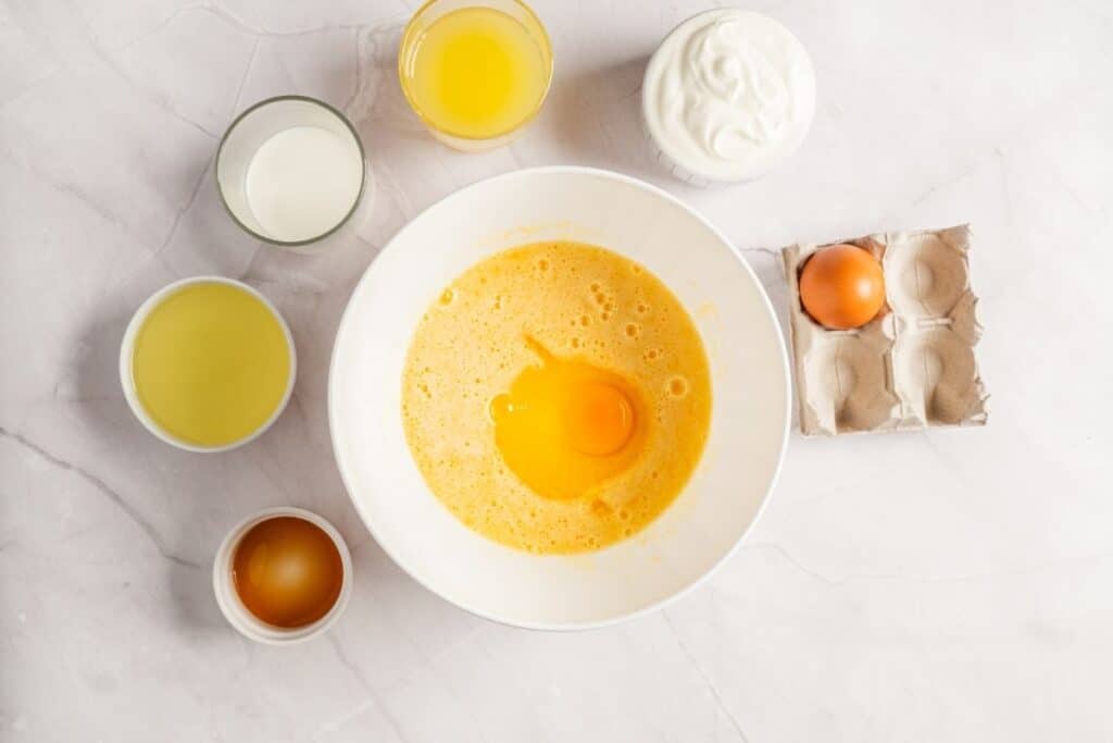 Overhead shot of a white bowl with whisked eggs, surrounded by other wet ingredients for a lemon loaf cake.