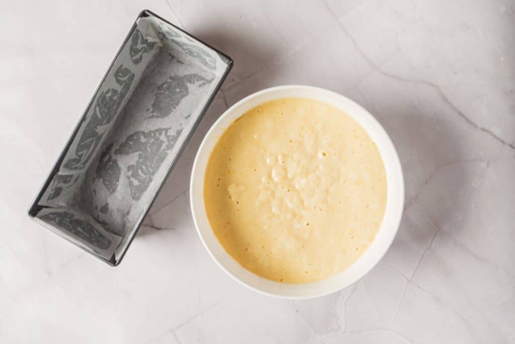 Overhead shot of a white bowl containing light yellow lemon loaf cake batter next to a parchment-lined loaf pan.