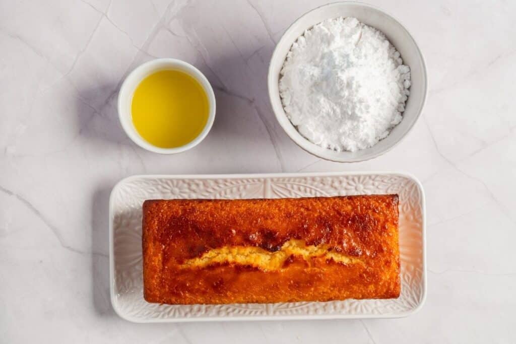 Overhead shot of a baked lemon loaf cake on a white plate, with a bowl of powdered sugar and a bowl of lemon juice.