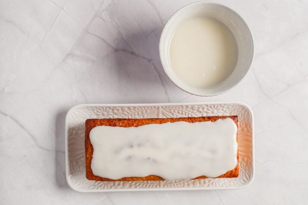 Overhead shot of a golden-brown lemon yogurt cake on a white plate, topped with a smooth white glaze, next to a bowl of more glaze.