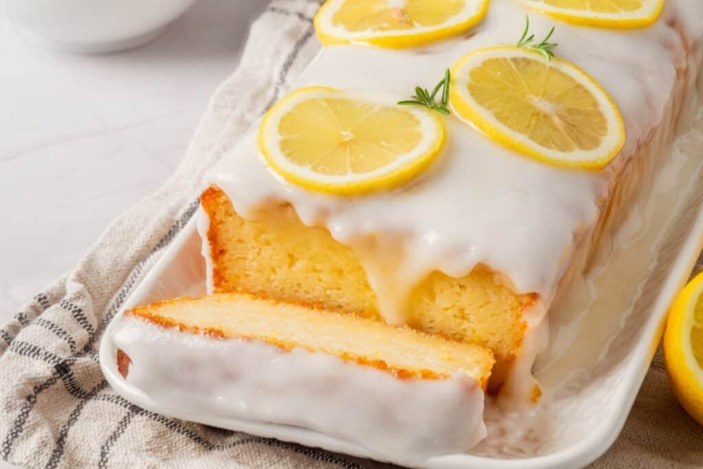 A close-up of a glazed lemon loaf cake with a slice partially cut, garnished with lemon slices and rosemary.