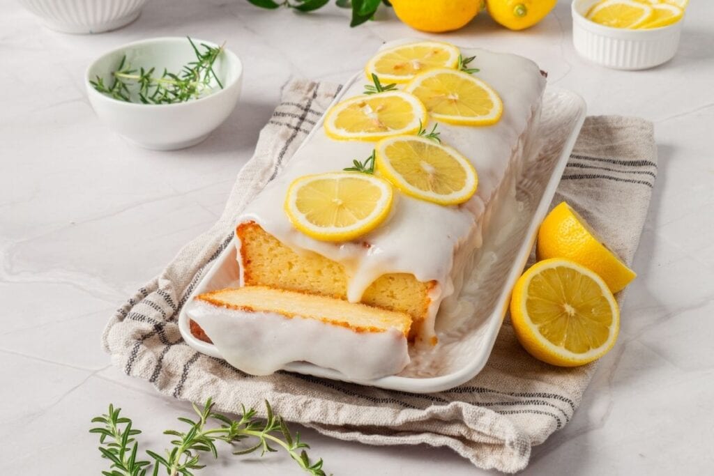 A glazed lemon loaf cake with a slice cut out, garnished with lemon slices and rosemary on a white plate.