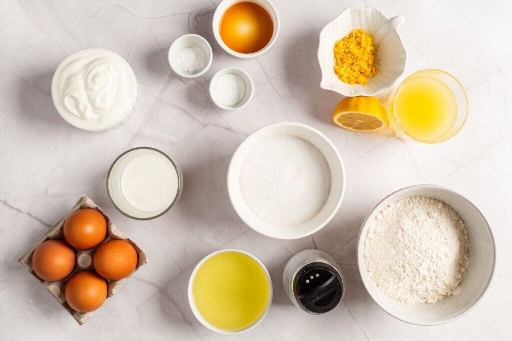 Overhead shot of labeled ingredients for a lemon loaf cake, including flour, sugar, eggs, yogurt, lemon juice, lemon zest, and baking powder.