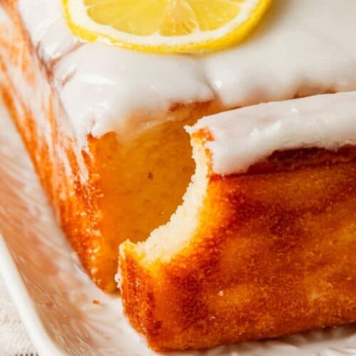 A close-up of a glazed lemon loaf cake on a white plate with a missing bite, revealing its light and airy texture.