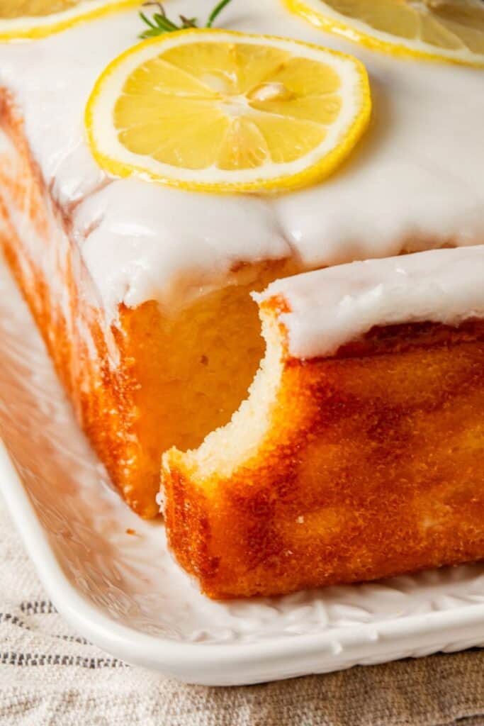 A close-up of a glazed lemon loaf cake on a white plate with a missing bite, revealing its light and airy texture.