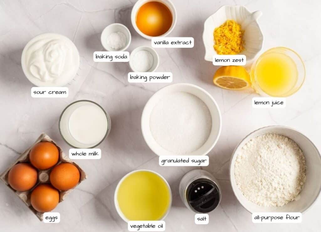 Overhead shot of labeled ingredients for a lemon loaf cake, including flour, sugar, eggs, sour cream, milk, lemon juice, lemon zest, oil, vanilla, baking soda, baking powder, and salt.