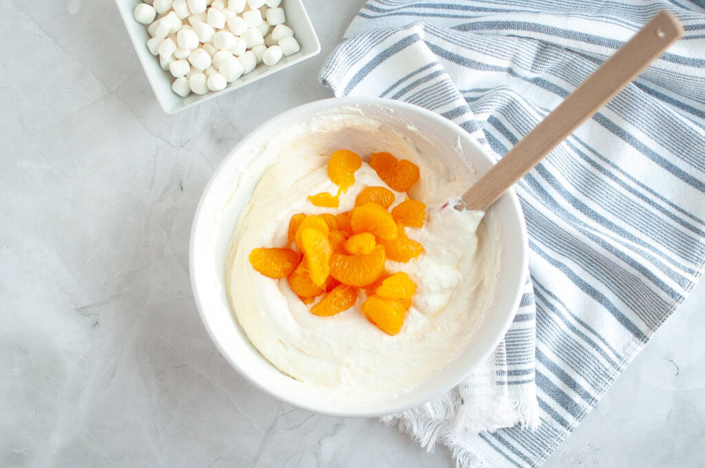 Overhead shot of bright orange mandarin orange segments being folded into a white, creamy mixture in a white bowl with a spatula.