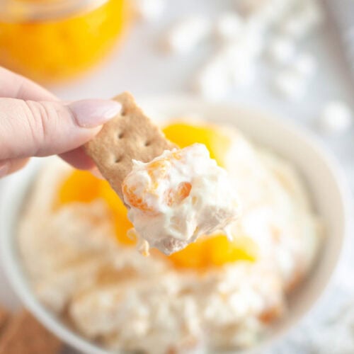 A hand holds a cracker topped with Orange Fluff Salad Dip over a bowl of the dip.