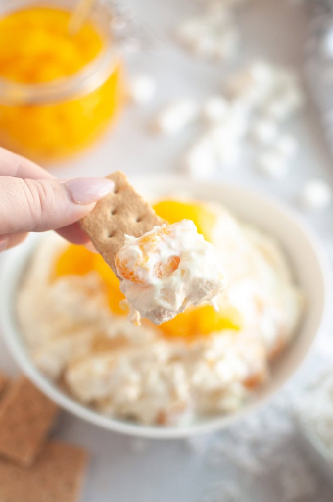 A hand holds a cracker topped with Orange Fluff Salad Dip over a bowl of the dip.