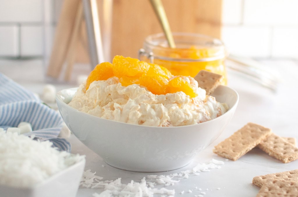 A white bowl filled with creamy Orange Fluff Salad topped with mandarin oranges and graham crackers.