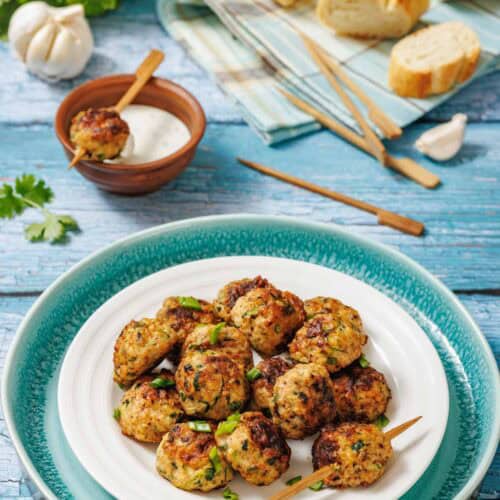 A plate of grilled meatballs garnished with herbs, served with a bowl of dipping sauce, skewers, and sliced bread on a rustic blue wooden table.