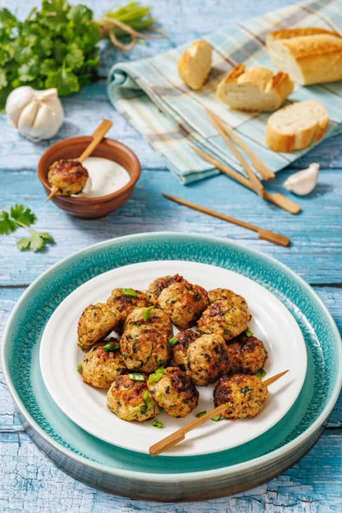 A plate of grilled meatballs garnished with herbs, served with a bowl of dipping sauce, skewers, and sliced bread on a rustic blue wooden table.
