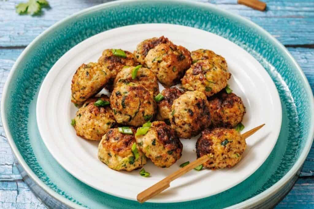 A white plate with grilled meatballs garnished with chopped green herbs, placed on a blue textured surface. A wooden skewer rests on the plate.
