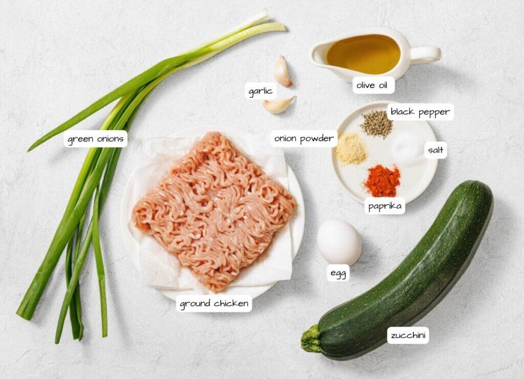 Ingredients on a white surface: ground chicken, zucchini, egg, green onions, garlic, olive oil, black pepper, salt, paprika, and onion powder.