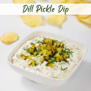 A bowl of creamy dill pickle dip topped with chopped pickles and fresh dill, with potato chips in the background.