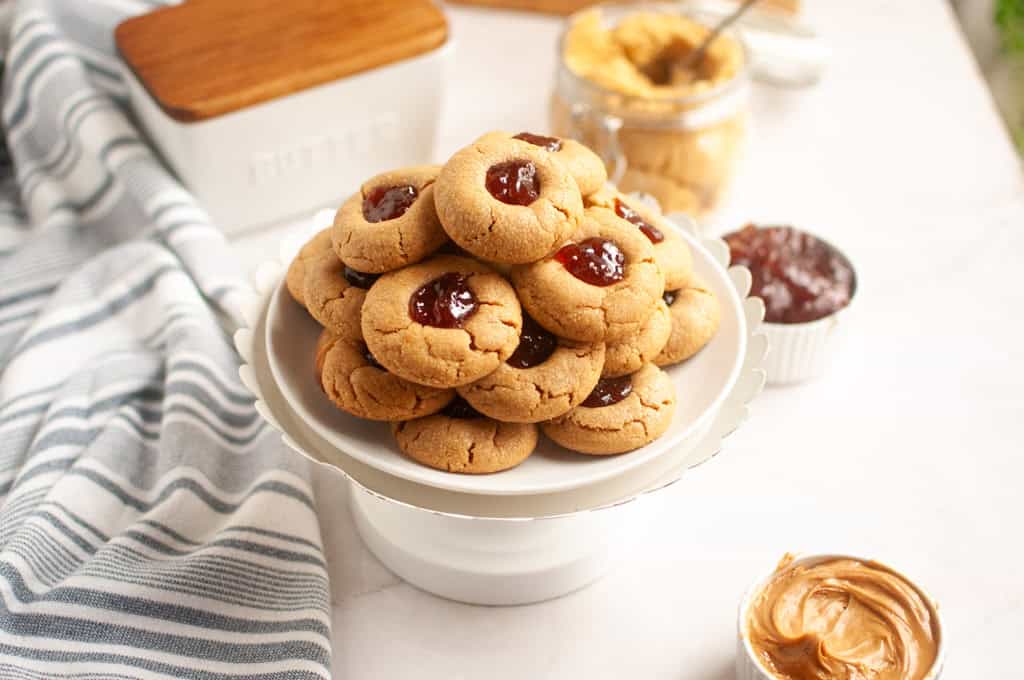 A stack of golden-brown peanut butter cookies with red jelly centers on a white pedestal plate.