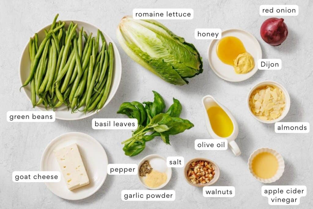 A variety of salad ingredients and seasonings, including green beans, romaine lettuce, red onion, cheese, nuts, basil, honey, Dijon, oil, vinegar, and spices, arranged on a white surface.