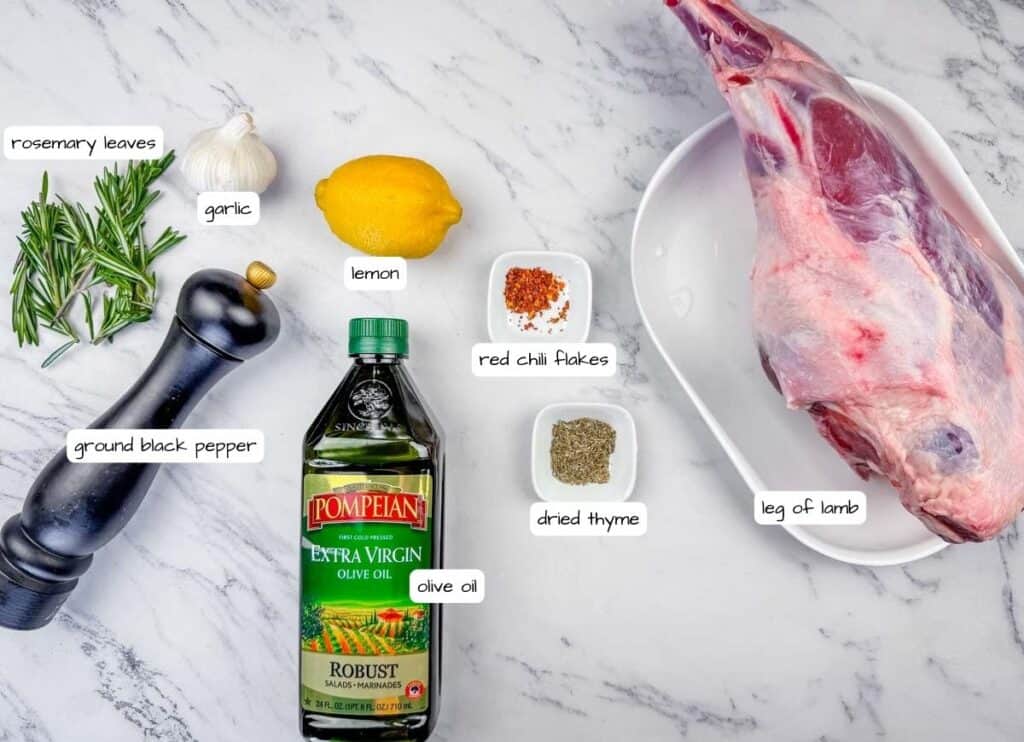 Overhead shot of labeled ingredients for roast leg of lamb, including a leg of lamb, Pompeian olive oil, lemon, garlic, rosemary, thyme, and red chili flakes.