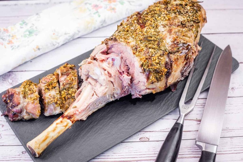A partially carved roast leg of lamb seasoned with herbs on a black serving platter with carving utensils.