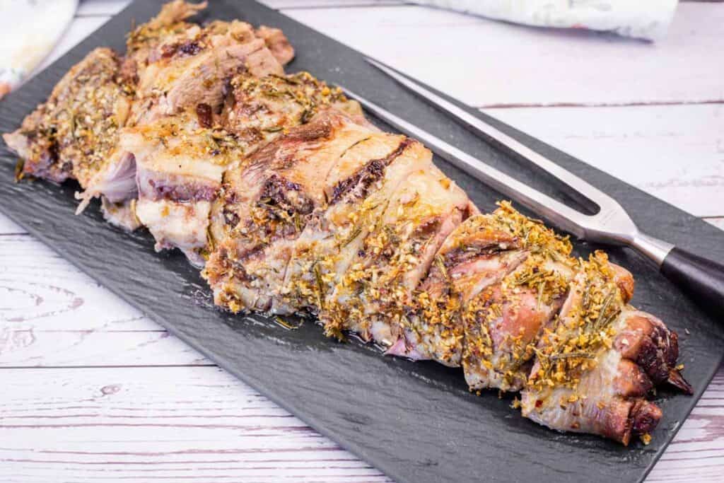 A black platter with sliced, roasted leg of lamb, seasoned with herbs.