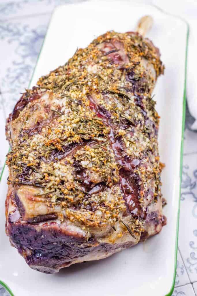 A roasted leg of lamb, seasoned with herbs, on a white platter.