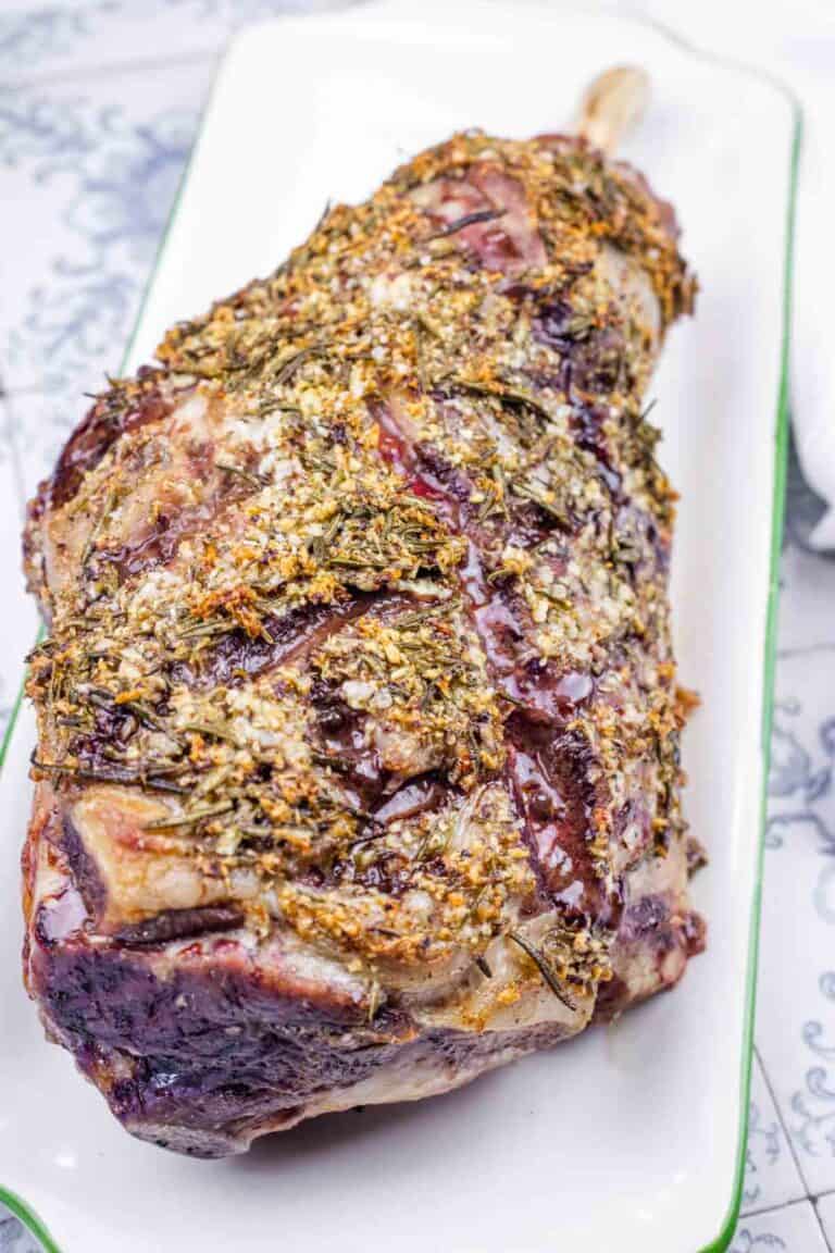 A roasted leg of lamb, seasoned with herbs, on a white platter.
