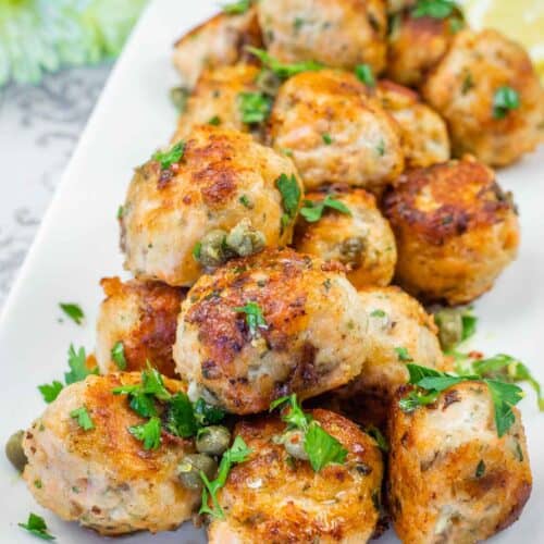 A pile of golden-brown salmon balls garnished with fresh parsley, capers, and lemon wedges on a white plate.