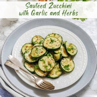 A white plate on a gray plate filled with rounds of sautéed zucchini, garlic, and herbs, with a fork and knife.
