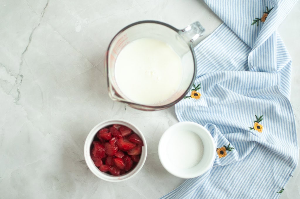 Overhead shot of sliced strawberries, milk (or buttermilk), and sugar on a marble surface with a blue and white cloth.