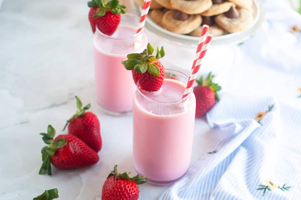 Two glasses of pink strawberry milk, garnished with fresh strawberries and red and white straws, on a white surface.