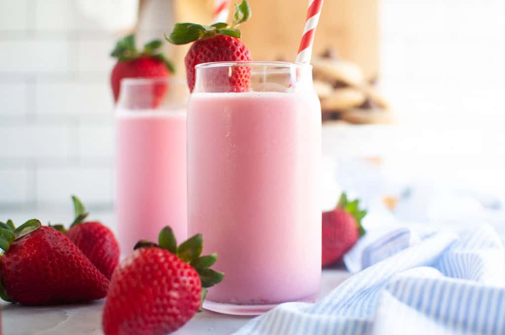 Two glasses filled with pink strawberry milk, garnished with fresh strawberries and red and white straws.