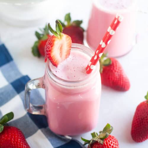 A pink strawberry milk in a glass jar with a red and white straw, surrounded by fresh strawberries.