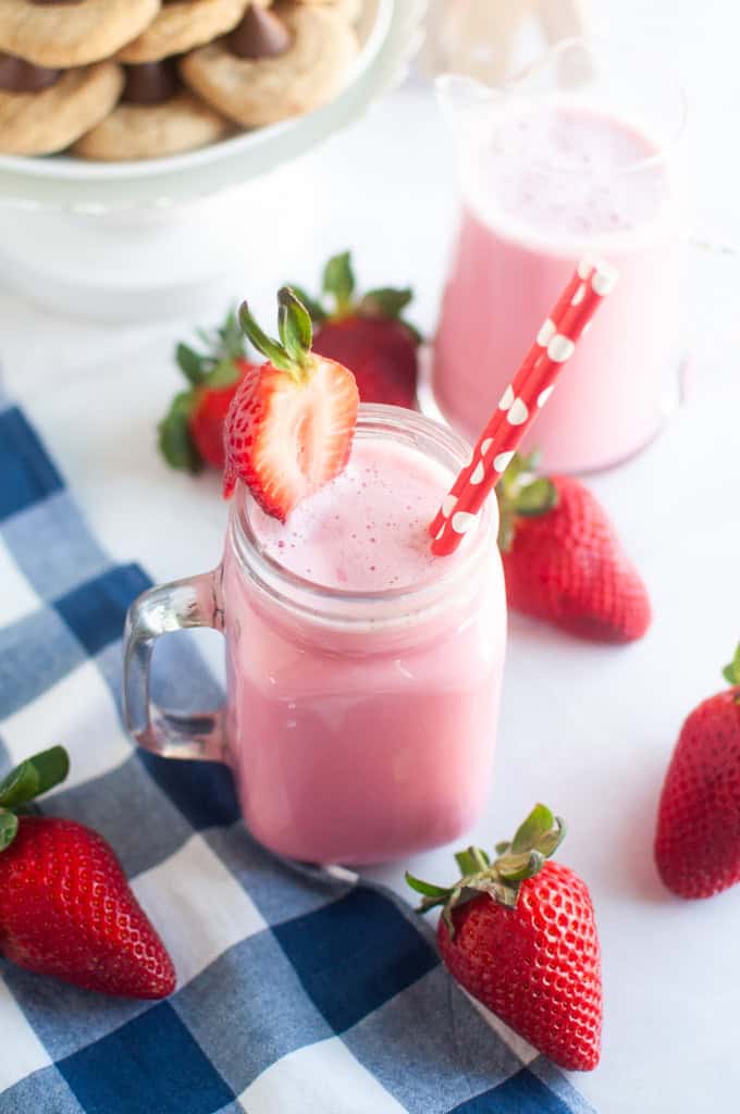 A pink strawberry milk in a glass jar with a red and white straw, surrounded by fresh strawberries.