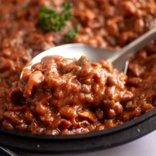 A close-up of a spoonful of baked beans in a thick, reddish-brown sauce, served in a black dish with a parsley garnish in the background.