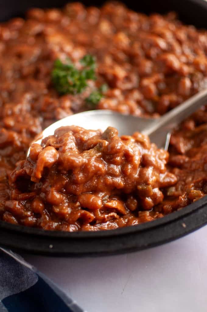 A close-up of a spoonful of baked beans in a thick, reddish-brown sauce, served in a black dish with a parsley garnish in the background.