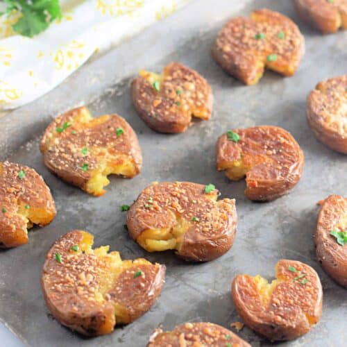 A baking sheet with several smashed, roasted potatoes topped with seasoning and herbs, with fresh parsley visible in the background.