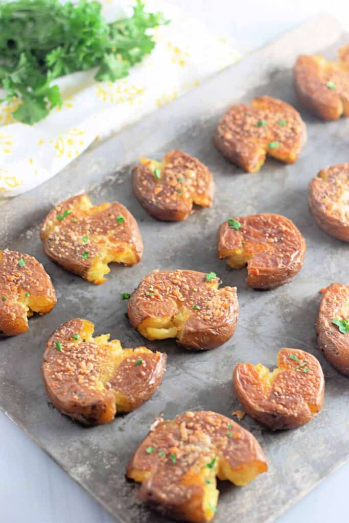 A baking sheet with several smashed, roasted potatoes topped with seasoning and herbs, with fresh parsley visible in the background.