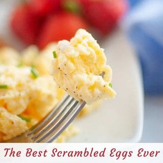 A fork holding a bite of scrambled eggs garnished with chives, with sliced strawberries in the blurred background. Text at the bottom reads, "The Best Scrambled Eggs Ever.