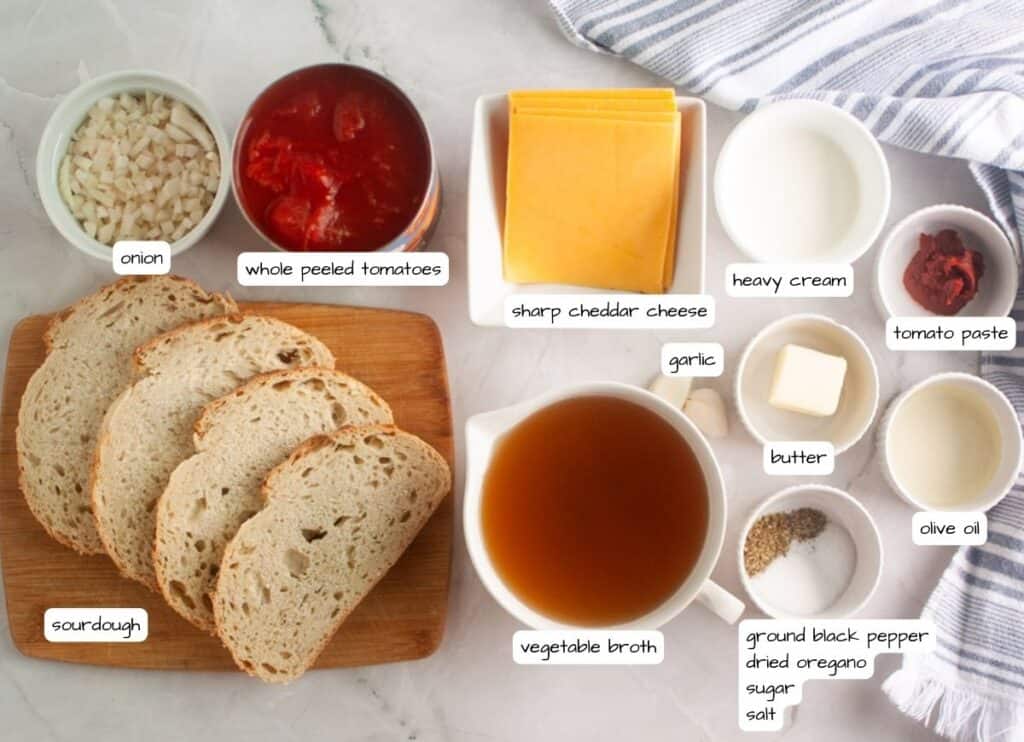 Ingredients for a soup are laid out, including sourdough bread, onion, whole peeled tomatoes, cheddar cheese, heavy cream, broth, and seasonings, all arranged on a white surface.