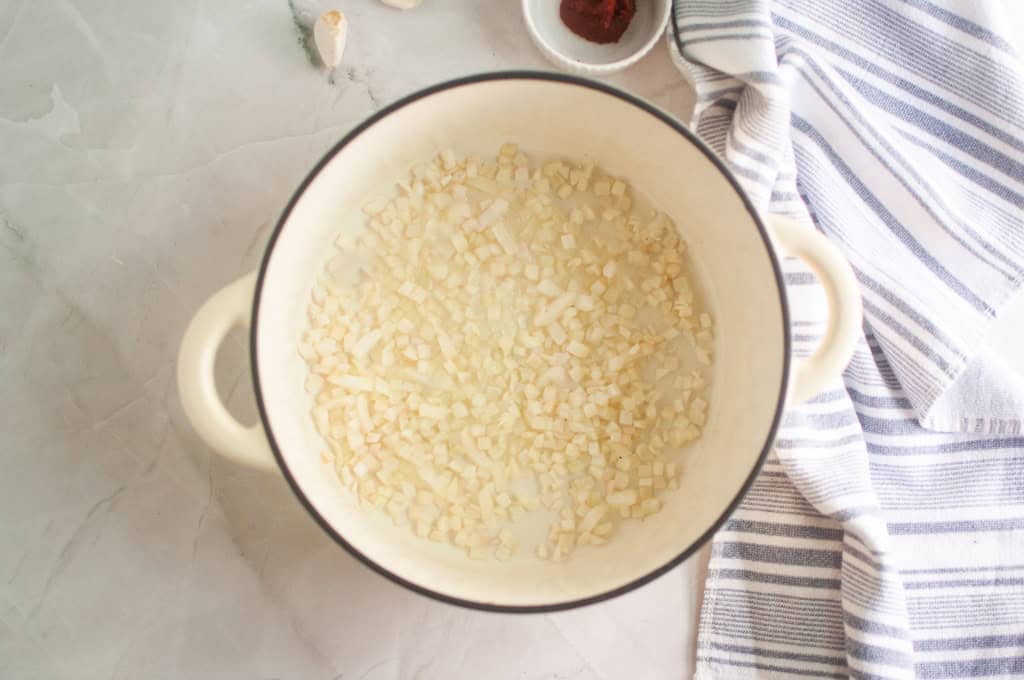 Diced onions are cooking in a white pot on a marble countertop, with a striped kitchen towel and small bowl nearby.