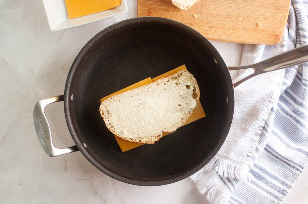 A slice of bread placed on top of a slice of cheddar cheese inside a black saucepan, with a cutting board and kitchen towel nearby.